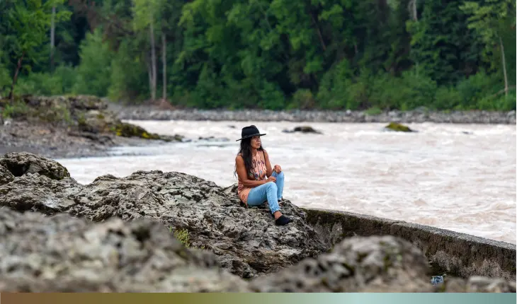 A woman seated on rocks beside a flowing river, surrounded by nature's tranquility and scenic beauty.